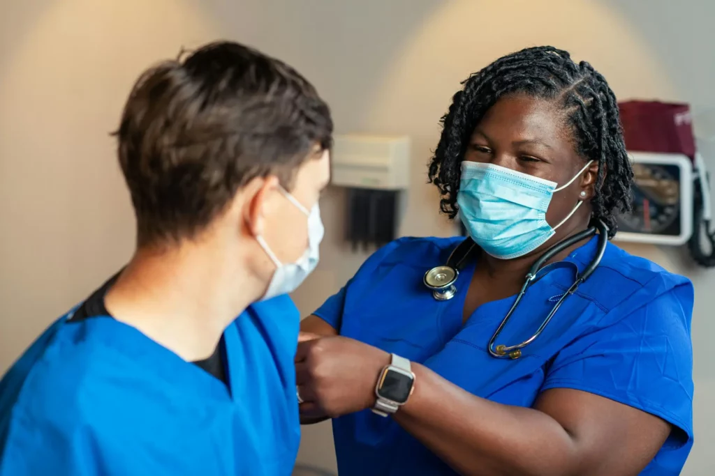 Healthcare worker in blue scrubs and face mask assisting a patient during a clinical care visit.
