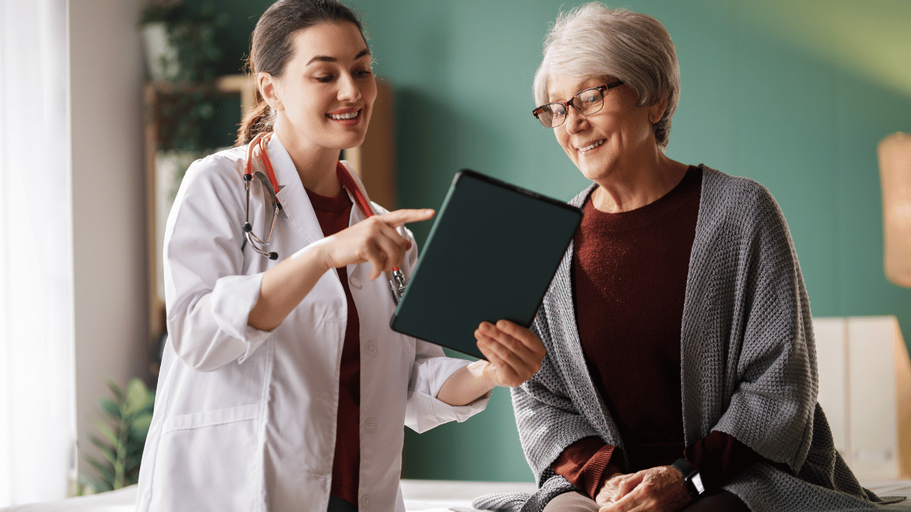 A friendly nurse in a white coat points to data on a dark handheld tablet while interacting with a smiling elderly resident at her bedside, demonstrating electronic Point-of-Care charting.