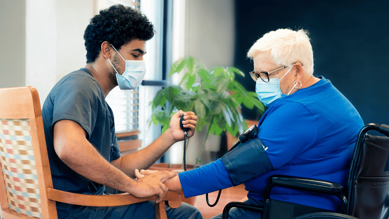 A masked male care provider in scrubs takes the manual blood pressure of a masked elderly woman in a wheelchair, illustrating a clinical data measurement task.
