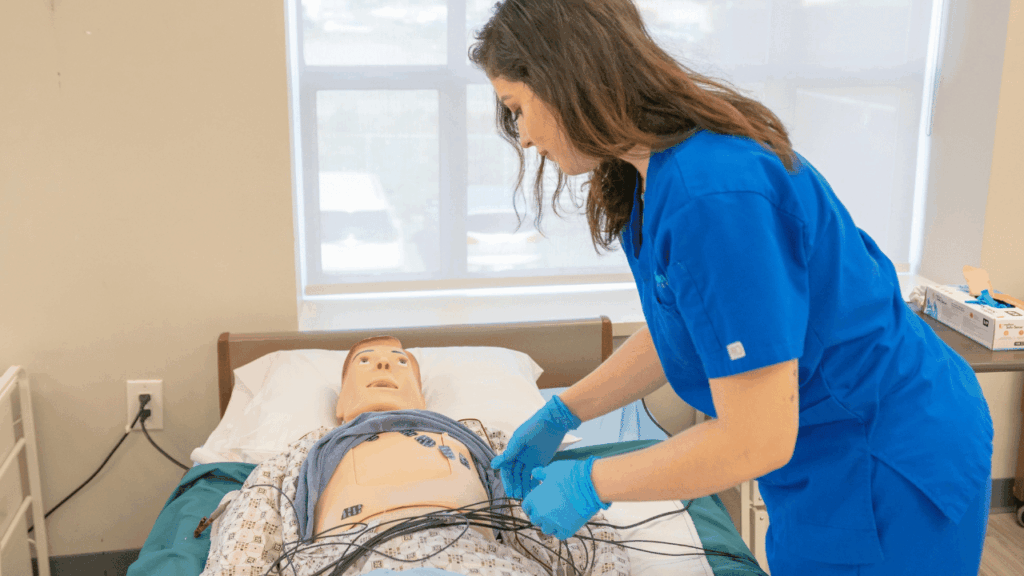 A focused nursing assistant student in blue scrubs and gloves practice clinical skills by attaching sensors to a simulation mannequin in a well-lit skills laboratory.