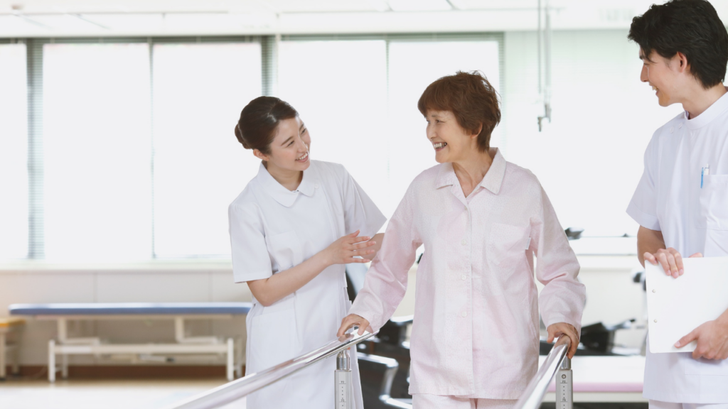 Two smiling CNAs provide close support and encouragement to an elderly resident walking along physical therapy parallel bars in a healthcare facility.