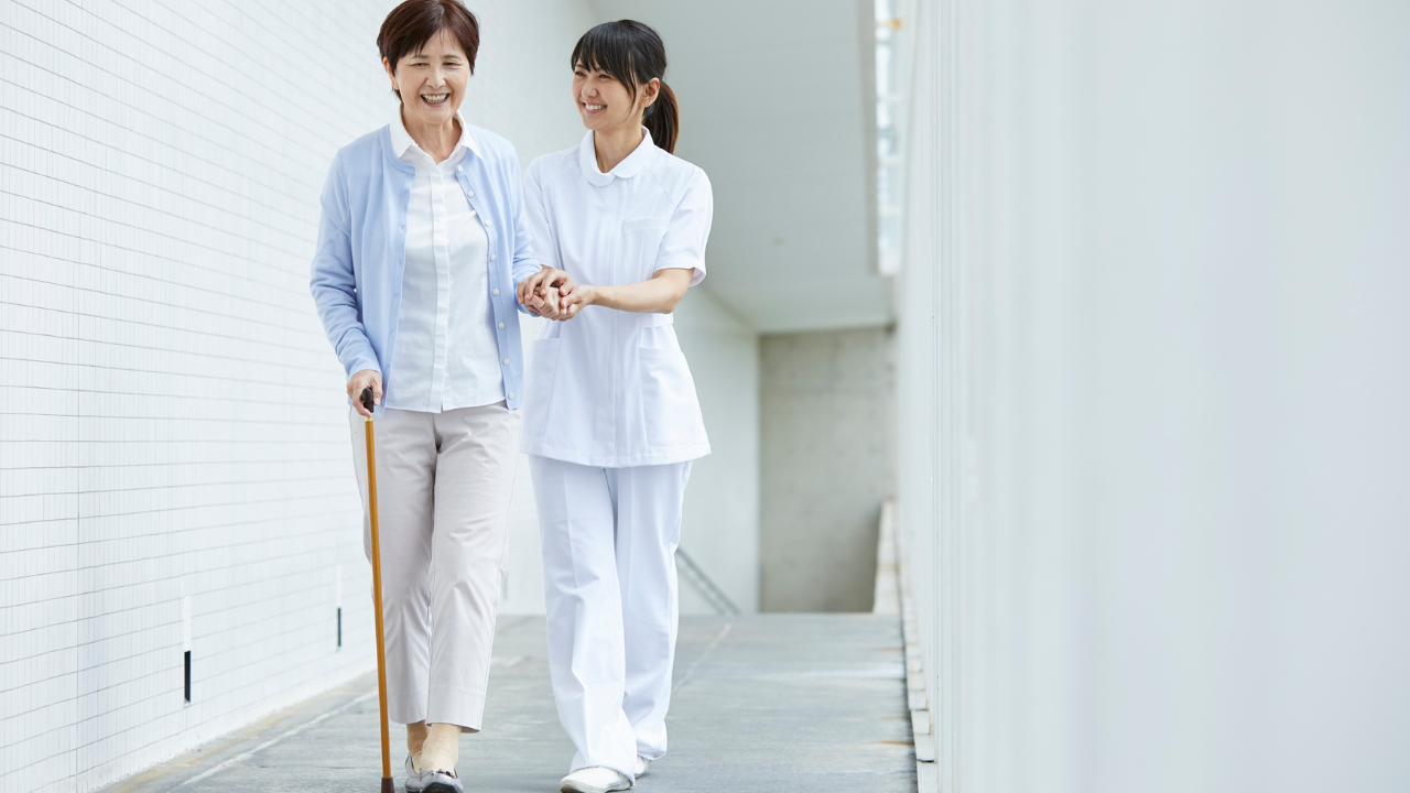 A smiling nurse holds the hand of an elderly woman using a cane while they walk together down a modern, white tiled corridor.