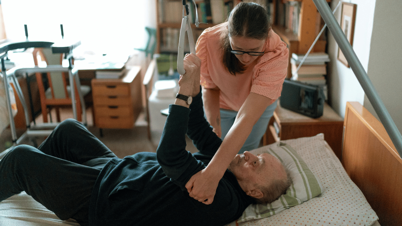 A home health aide assisting an elderly man to sit up in his bed by holding onto his arm and the overhead pull handle for support.