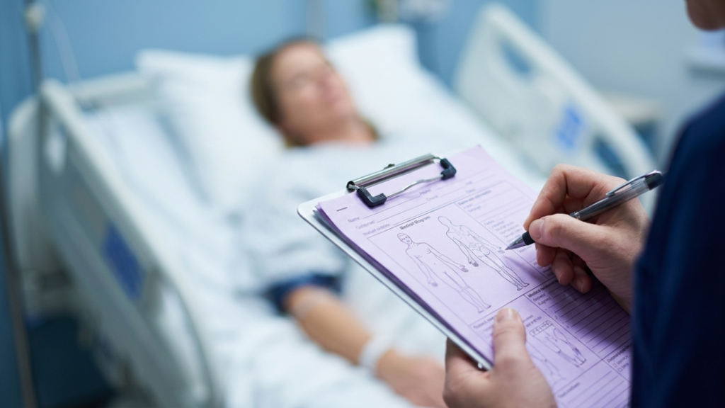 A nurse with a stethoscope documents a patient's condition on a purple charting form with anatomical diagrams at the bedside.