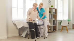 CNA in blue scrubs assisting an elderly woman in using a walker for mobility.