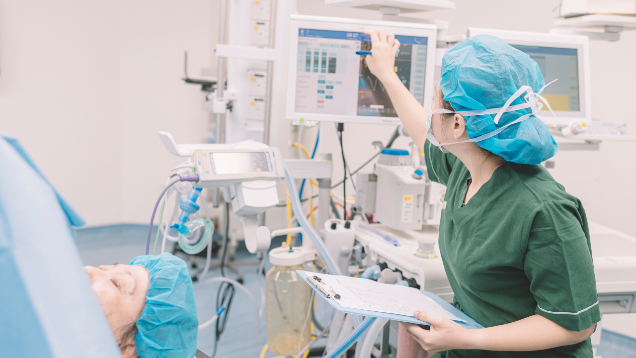 An East Asian female CRNA intently monitors vital sign waveforms and adjusts anesthesia controls during a surgical procedure.