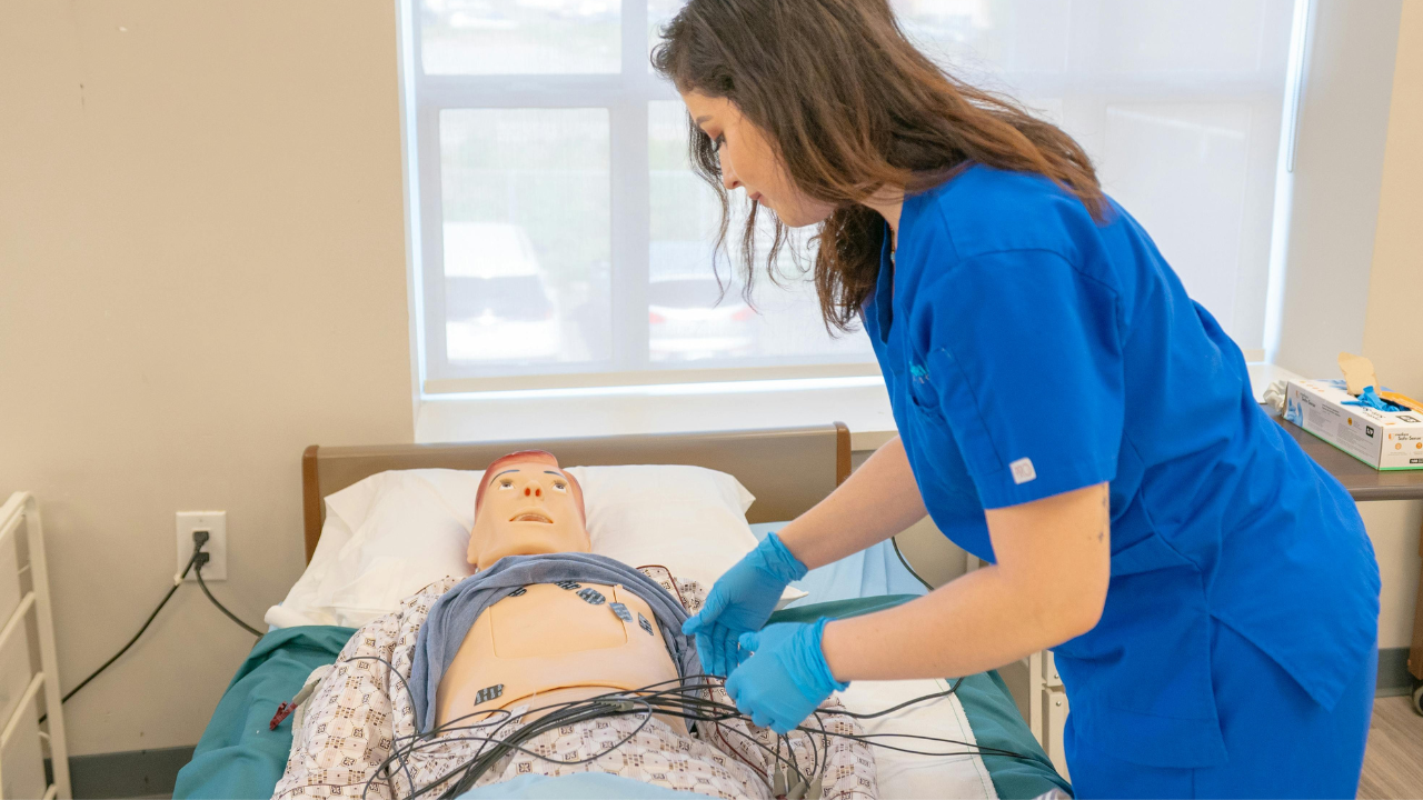 A female nursing student in blue scrubs and gloves practices connecting leads to a patient simulator mannequin in a lab.